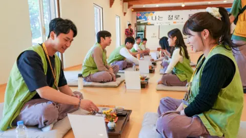 A row of men sitting and a row of women sitting on the floor of a temple across from each other