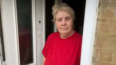 Patricia Foulds standing at her front door looking at the camera. She has short hair and is wearing a red T-shirt.