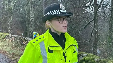 The police officer, who is wearing a police hat, fluorescent green jacket and a pair of glasses, speaks while standing near a mossy, stone wall and line of trees.