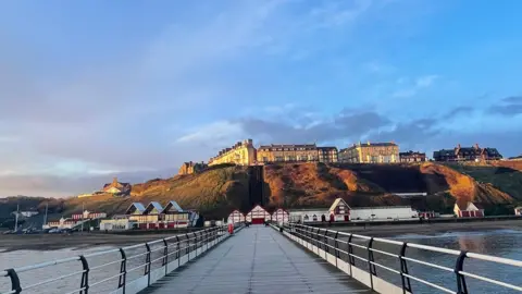 Sarah n Essie A view of Saltburn-by-the-Sea looking back along its pier. Victorian houses top a high grassy bank, at the foot of which red and white heritage buildings, including beach huts, are spread along the sea shore.