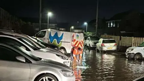 Picture shows flooding in a residential street. Welsh water engineer stands in front of a Welsh Water van working to sort the problem. 