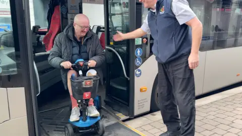 Eric Corkish is on a blue mobility scooter in the doorway of a silver bus. He is on a ramp resting on the pavement and the bus driver is to the right, with a hand out to help. They are both smiling.