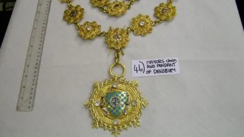 Highly decorated gold chain and pendant on a white tablecloth, with a transparent ruler next to it