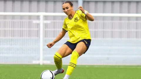 Lucy Copsey Connolly-Brame is wearing the yellow and blue Hashtag United kit, as she goes to strike a white football with her right foot. It is during a game..