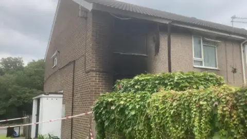 An end-of-terrace property has fire damage around a first floor window. Police tape surrounds the building.