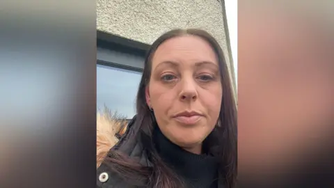 Lesley Bahreini. She has long dark hair with a middle parting and green eyes. She is standing outside a building with pebbledash an a window with a black frame. She is looking at the camera with a serious expression and she is wearing a black puffer jacket with faux fur around the hood.