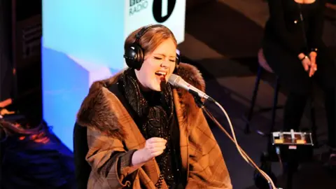 Adele performing in the Live Lounge in 2011, wearing a brown overcoat, black headphones and singing into a silver microphone