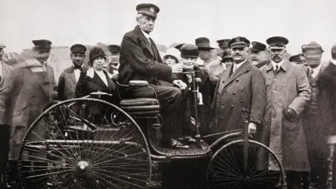 A black and white photo of Karl Benz displaying the Benz Patent-Motorwagen to a crowd of people. He is sat high up in the vehicle, which has large back wheels and a smaller front wheel, and is donning a suit and hat. 