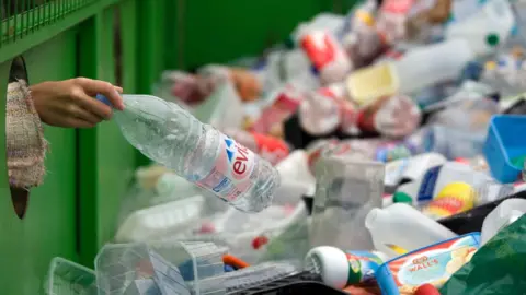 Someone's hand poking through into a skip holding a plastic bottle.