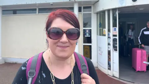 A woman standing outside the airport arrivals building. They are wearing sunglasses, earrings, a chunky chain-link necklace, and a backpack. In the background, people are exiting through automatic sliding doors, including one person pulling a pink suitcase.