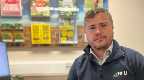 Aled Davies, wearing a blue NFU Cymru branded fleece, looks at the camera while sitting at a desk in an office