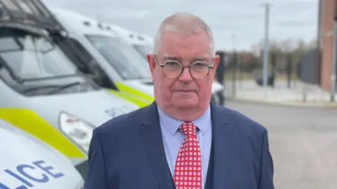 Laura Foster/BBC John Tizard with very short grey hair and round silver-framed glasses wearing a dark blue jacket, red and white tie and blue shirt, standings in front of some white and yellow police cars and vans. There is a paved area behind.