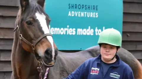 A man wearing a blue jumper and trousers is stood infront of a brown and white horse holding a trophy in the shape of a horse's head. He is wearing a green helmet.