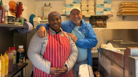 Violet John and James Nas, the owners of the Hot Sausage Company, standing in their van. They are both smiling into the camera. Violet is wearing a red and white apron and James is wearing a blue jacket.