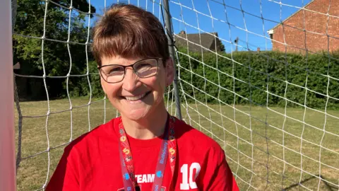 Henry Godfrey-Evans/BBC A woman with a red top on and glasses smiling at the camera while stood in front of a goal