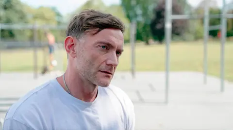 Christian is standing in front of gym equipment looking off to his right. He looks serious as though he is listening intently to something being said. He is a white man with blue eyes, brown hair and has stubble. He is wearing a light blue tee shirt and a silver necklace