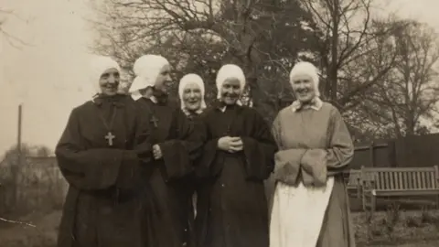 Five women stand together, facing the camera and smiling in a sepia-toned photograph. Behind them is a large tree with a wooden bench beneath it. Four of the women are wearing the black habit clothing of nuns, with white headpieces. The woman on the far left has a large crucifix hanging from her neck in front of her clothes. The woman on the far right is wearing a lighter coloured robe with a white pinafore tied around her waist and a white headpiece.
