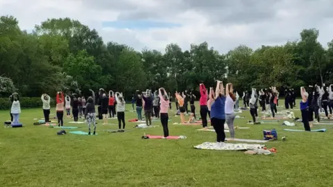 A large group of people standing up doing a yoga pose. They are on mats or rugs and are wearing a different coloured tops and trousers or leggings, which are mainly black. The group are outside in a grassy area. 