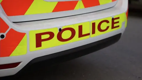 BBC An image of the rear of a marked police car. The image shows the car's rear bumper. The vehicle is white and has POLICE written in dark red on a luminous yellow background