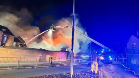 A aerial fire ladder hosing down water from 20 feet in the air. Two firefighters are in vision.