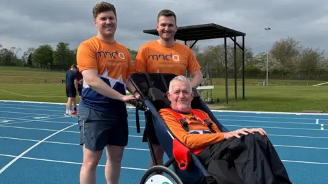 Two men stand on a running track next to a man in a wheelchair - all are wearing orange MND Association T-shirts.