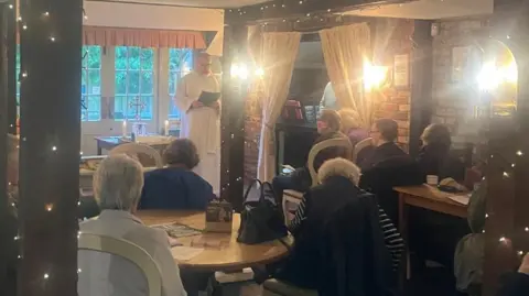 A group of worshipers in a pub. There is a priest stood speaking to the group. 