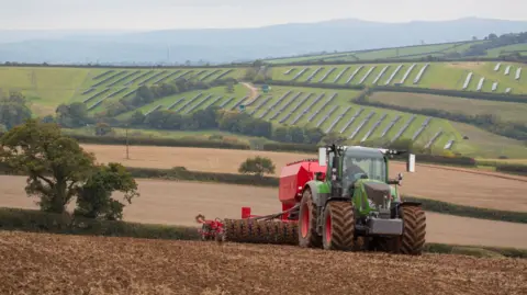 Getty Images A green and red tractor is pulling a cultivator and drill across a muddy field. On the hills in the background lie solar panels. A light mist sits in the sky.
