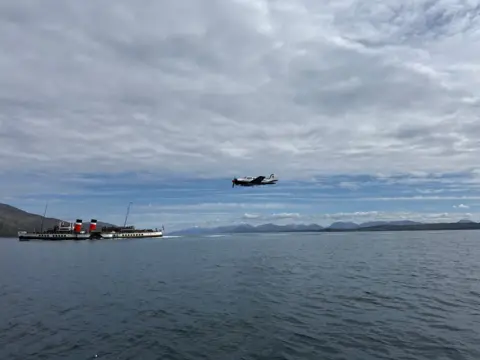 Raymond Codona Passenger steamship with two red-and-black funnels sailing across calm water, while a small propeller plane flies overhead beneath a wide sky filled with low clouds and distant mountains