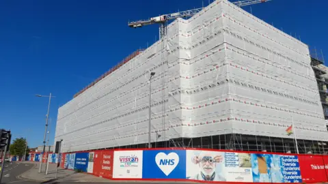 South Tyneside and Sunderland Trust The building that will host Sunderland’s new eye hospital is covered in protective grey sheeting. NHS boards are all around the lower floor of the site. A crane is visible above the building.