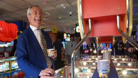 Joe Giddens/PA Nigel Farage standing next to a game in an amusement arcade. He is wearing a deep blue jacket, white shirt and patterned tie with what look to be butterflies on it. He is holding a cup, which presumably contains coins. There are other people in the background who are out of focus. 