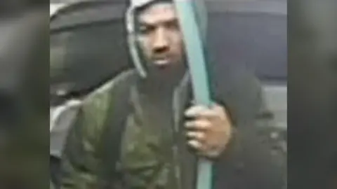 A man wearing a brown coat with a grey hood on and a black rucksack on.