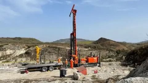 Cornish Lithium Mining A drilling rig clad in orange coloured external metalwork sits atop a landscape which appears to show a history of mineral extraction evidenced by the hinterland of large pits and piles of what appear to be waste material, alongside three people attired in orange clothing and wearing hard hats.