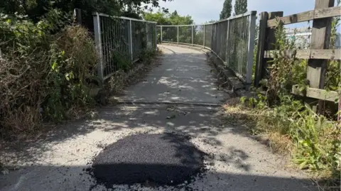 Phillip Norton / BBC An entrance to a footpath, with railings on either side. There is a circle of black ashfalt in front of the path where a concrete bollard was. 
