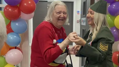 An elderly woman dressed in a red jumper with the image of a penguin on it is smiling holding hands with a younger woman who is wearing a fancy dress army uniform. She has long blond hair and is smiling at the older lady. They are both surrounded by balloons.