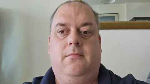 Peter Clarke Peter Clarke in a selfie. He has short hair and is wearing a polo top. He appears to be in a domestic setting like a living room or lounge.