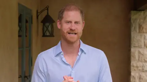 Prince Harry in a light purple shirt in front of a stone wall