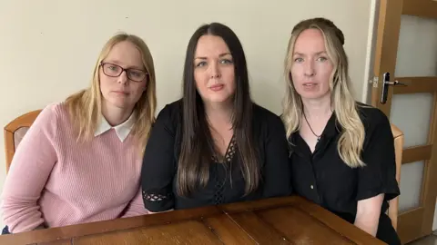 Three women sat at a kitchen table in a row. They are looking at the camera and have serious looks on their faces.