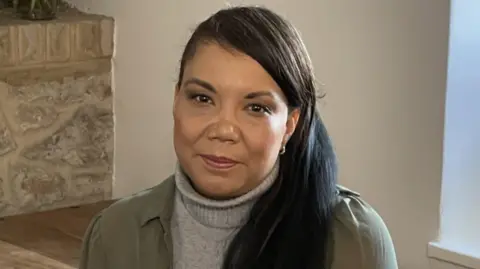 Caz Goddard is looking at the camera. She has long dark hair in a side ponytail and is wearing a khaki shirt over a grey knitted roll neck.