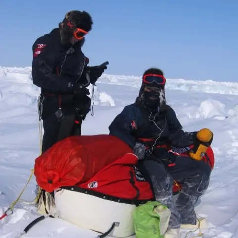 Karl Bushby Two men, both in extreme weather clothing, on the frozen Bering Strait. One of the men is standing while the other sits on a sledge containing their tent and other provisions.