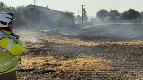 A firefighter wearing a hi-vis jacket and white helmet looks on at the charred field, which has smoke rising from it. There is a large agricultural building to the left and there are trees around the field's perimeter.