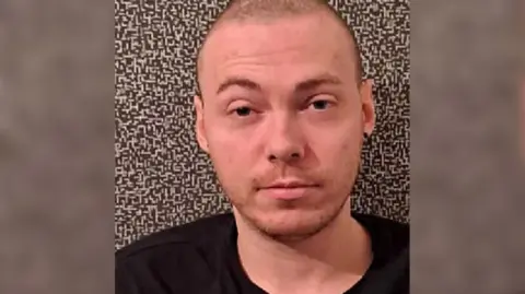 Grant McKinnon is looking straight at the camera. He has a shaved head and is wearing a black top