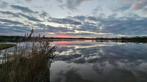scuddingcloud A cloudy sky is reflected in a lake. There is an orange tinge on part of the horizon.