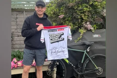 Joe Dick A man in a black hoodie, grey shorts and black cap stands by a bike in a garden holding up a white cycling T-shirt which says Fitness Worx and Campaign Against Living Miserably
