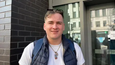 Calumn Donaghey, 30, who has dark blonde hair, a pair of sunglasses on his head and is wearing a dark blue gilet over a white t-shirt. He is standing outside a hotel doorway smiling at the camera