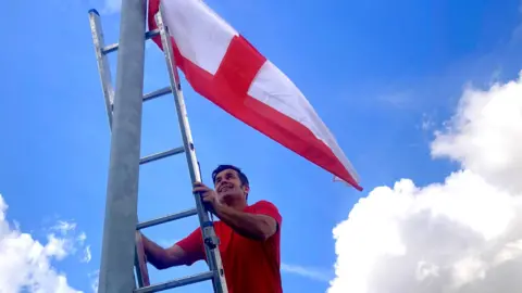 A man wearing a red T-shirt climbing a ladder to fix a St George's flag to a lamp-post.