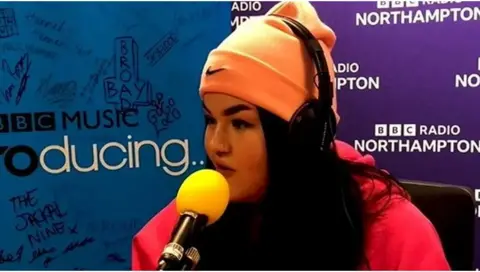 Mae Stephens is wearing an orange Nike beanie hat and a pink hoodie. She is speaking into a studio microphone and has BBC Music Introducing and BBC Radio Northampton branding behind her.