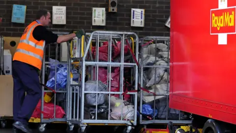 John Giles/PA Wire A Royal Mail worker is pushing a large metal trolley with piles of post in it. 