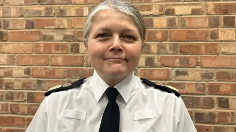 Chief Constable Sarah Crew standing looking at the camera. She is wearing a white shirt and black tie