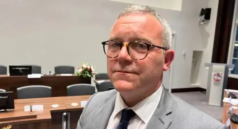 Simon Dedman/BBC Will Russell looking side-on at the camera in a council chamber. He has short grey hair and leopard-print round glasses, and is wearing a white shirt, navy tie and a grey jacket.