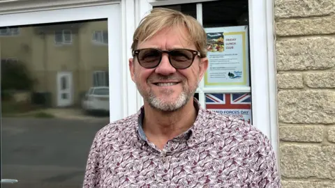 A photo of Andrew Morris-Wyatt, from Chalford Parish Council. He is white with light brown hair and a beard and is wearing glasses. He is stood in front of a building 